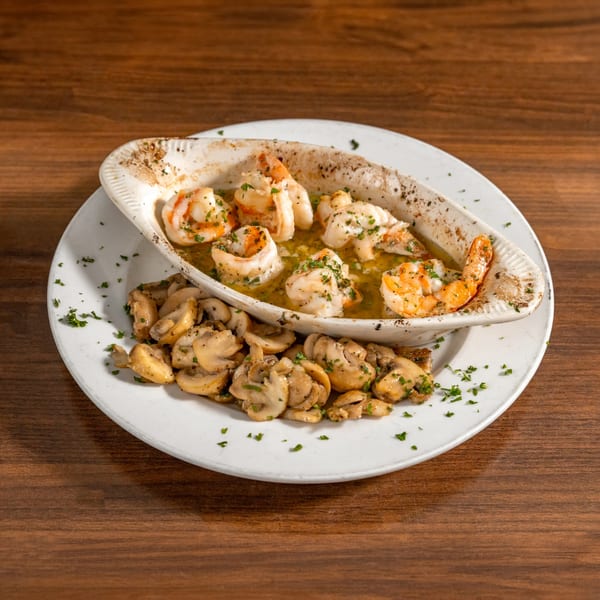 Shrimp scampi in a white dish with sautéed mushrooms on a white plate, on a wooden table.