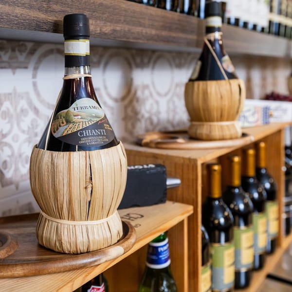 A Chianti wine bottle in a straw basket, with other wine bottles on wooden shelves.