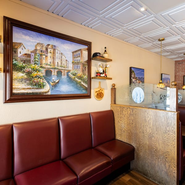 Cozy Italian restaurant interior with red booth seating, framed canal painting, and decorative shelves.