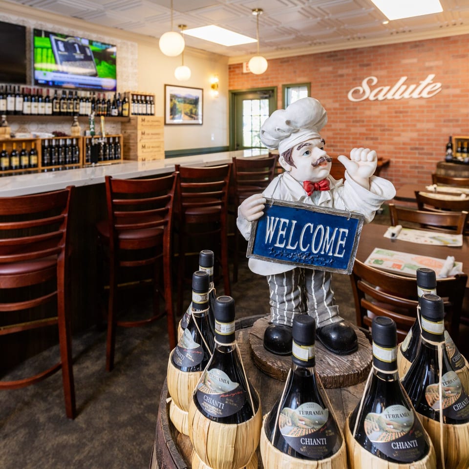 A chef statue holding a "Welcome" sign, surrounded by Chianti bottles, in an Italian restaurant.
