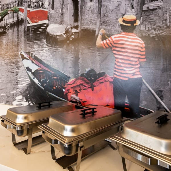 Three chafing dishes on a counter with a mural of a gondolier in Venice behind them.