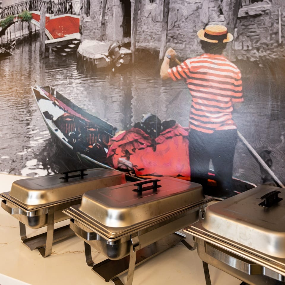 Three chafing dishes on a counter with a mural of a gondolier in Venice behind them.