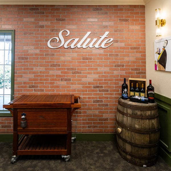 A brick wall with a "Salute" sign, a wooden cooler, and wine bottles on a barrel.