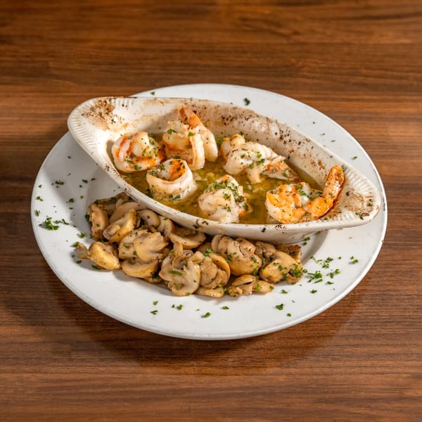 Shrimp scampi in a white dish with sautéed mushrooms on a white plate, on a wooden table.