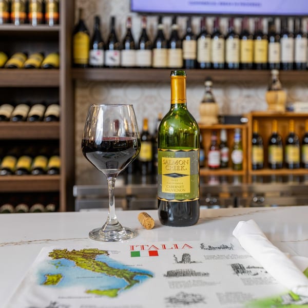 A glass of red wine and a bottle of Salmon Creek Cabernet Sauvignon on an Italian-themed placemat.
